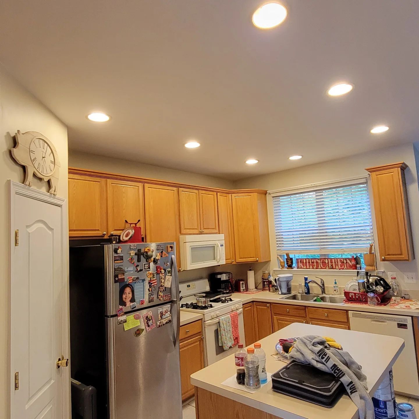 Kitchen with wood cabinets, stainless fridge, white stove, window blinds, and a cluttered island countertop.
