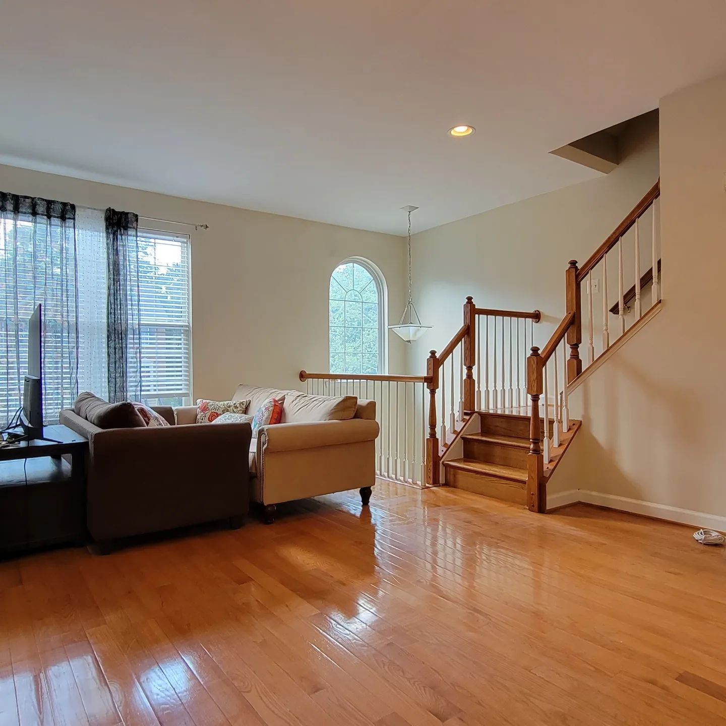 Empty living room with hardwood floors, beige sectional, large window, and staircase with wooden railing