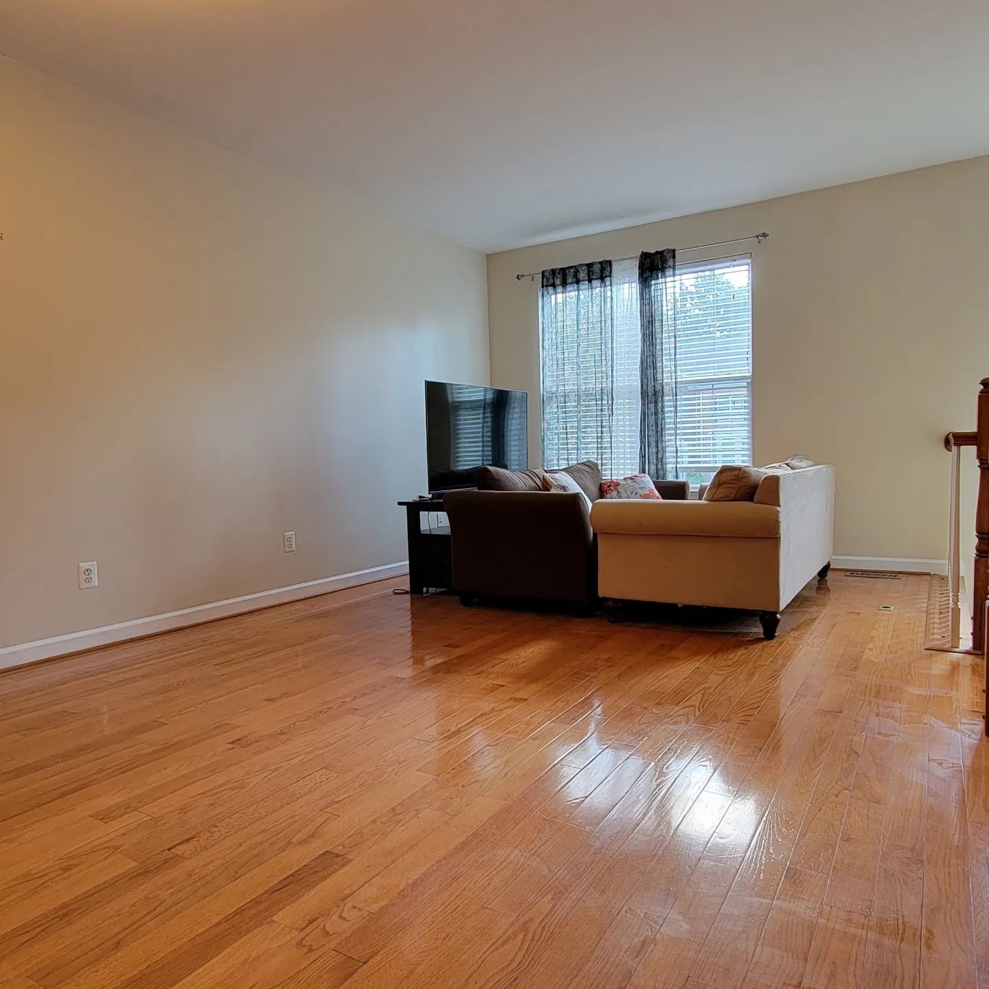 Empty living room with hardwood floors, beige walls, a sofa, TV, and sunlight through a window.