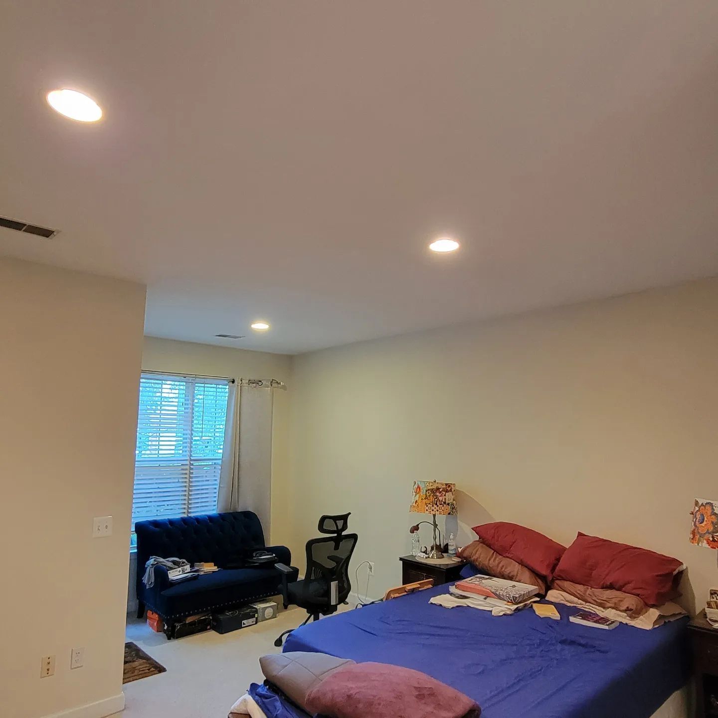 Bedroom with blue bedspread, red pillows, dark chair, and window with curtains in a softly lit room