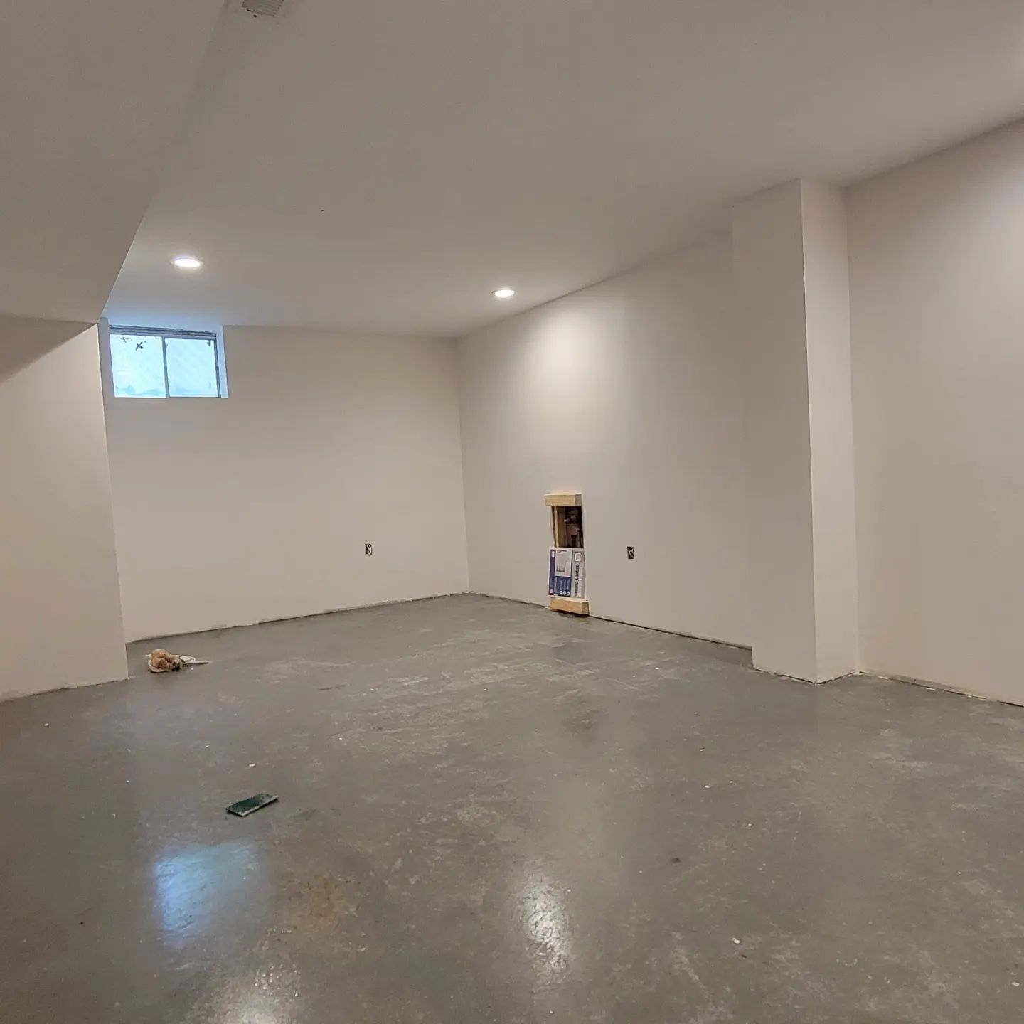 Empty unfinished room with white walls, concrete floor, and a small window near the ceiling.