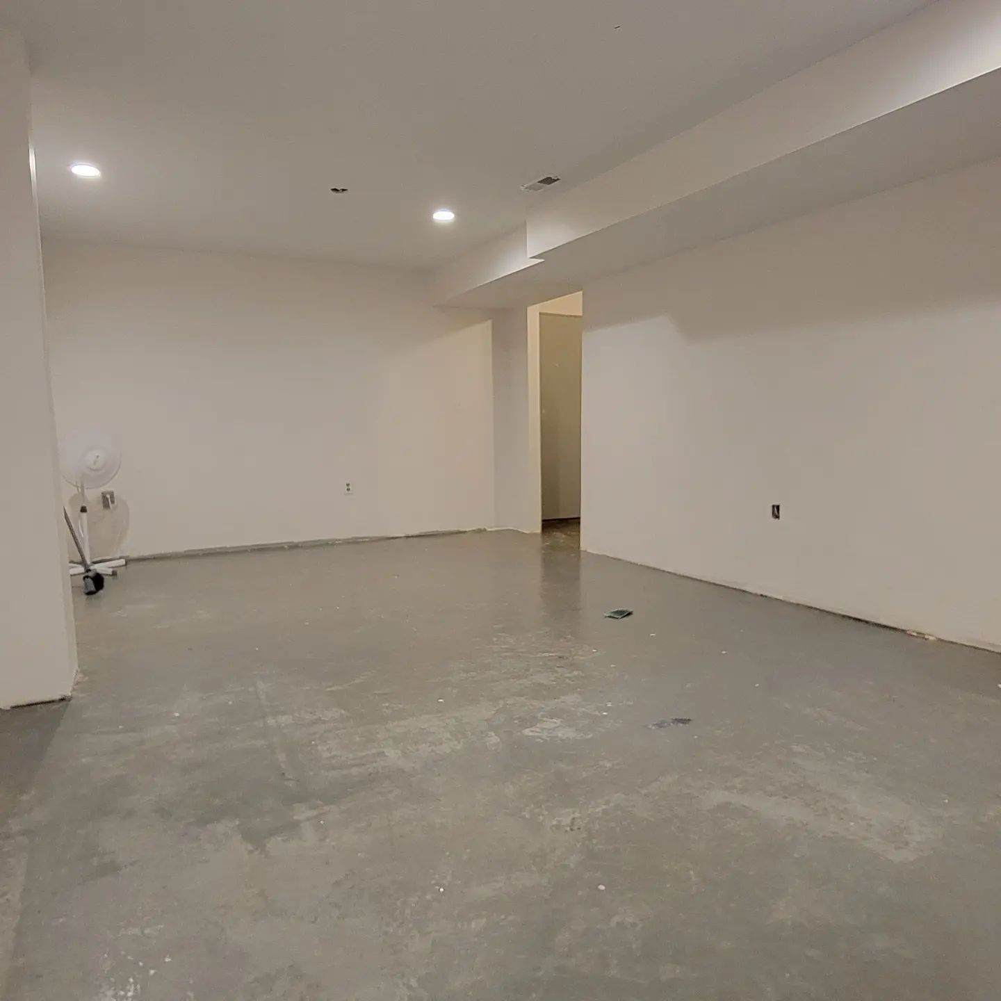 Empty unfinished room with concrete floor, white walls, recessed lights, and a doorway opening to another space