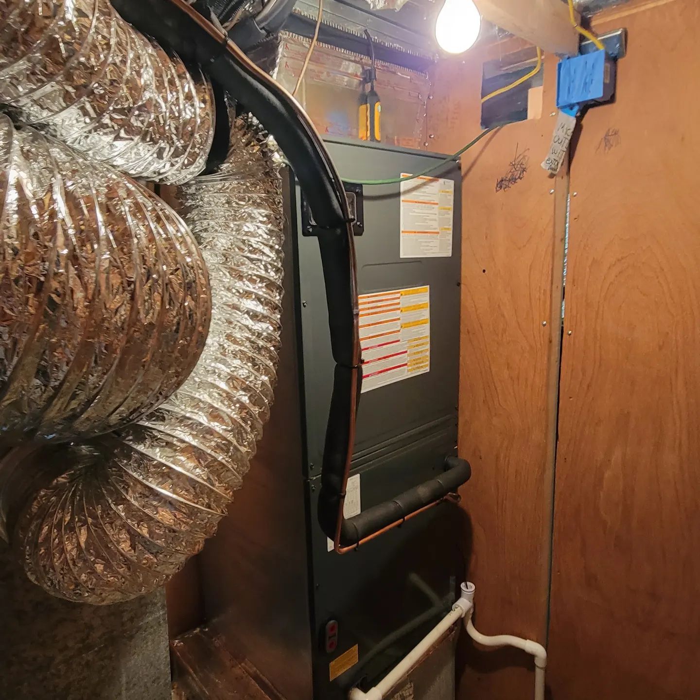 Basement HVAC unit with ductwork, exposed pipes, and a hanging light bulb against wood-paneled walls