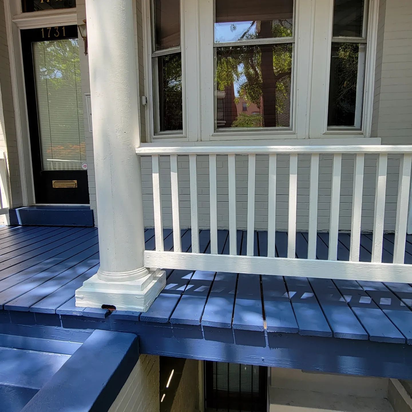 Blue porch with white railings and columns outside a house, with windows and a front door.