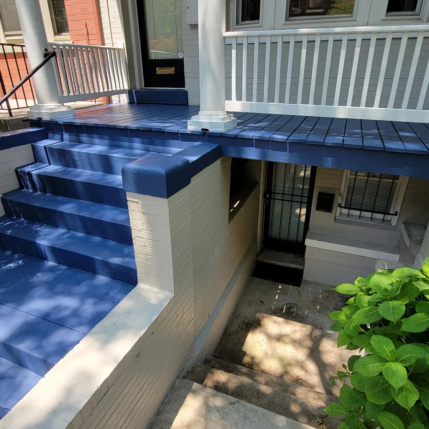 Blue-painted front porch with stairs and railing above a lower entryway and shaded walkway