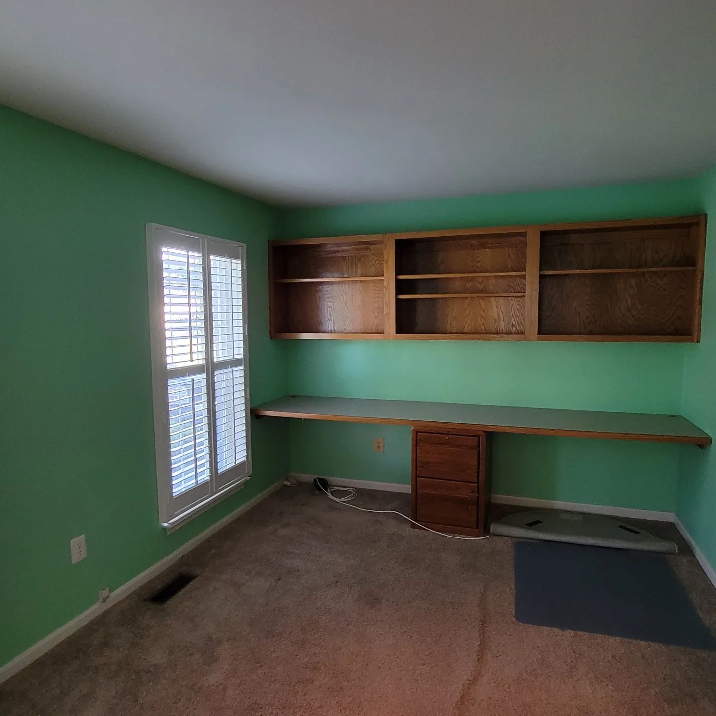 Empty room with green walls, built-in wooden shelves, window blinds, and a desk along the back wall