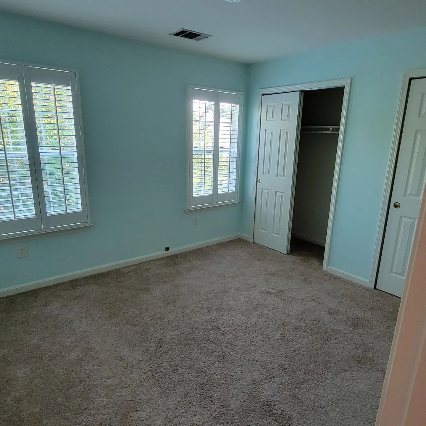 Empty light blue bedroom with beige carpet, white shutters, and an open closet door