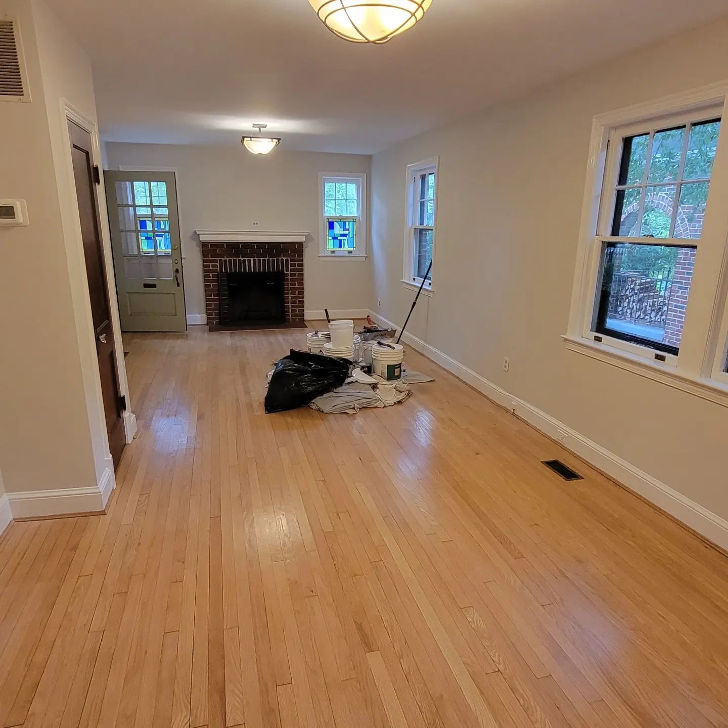 Empty living room with light wood floors, fireplace, windows, and moving boxes near the center.