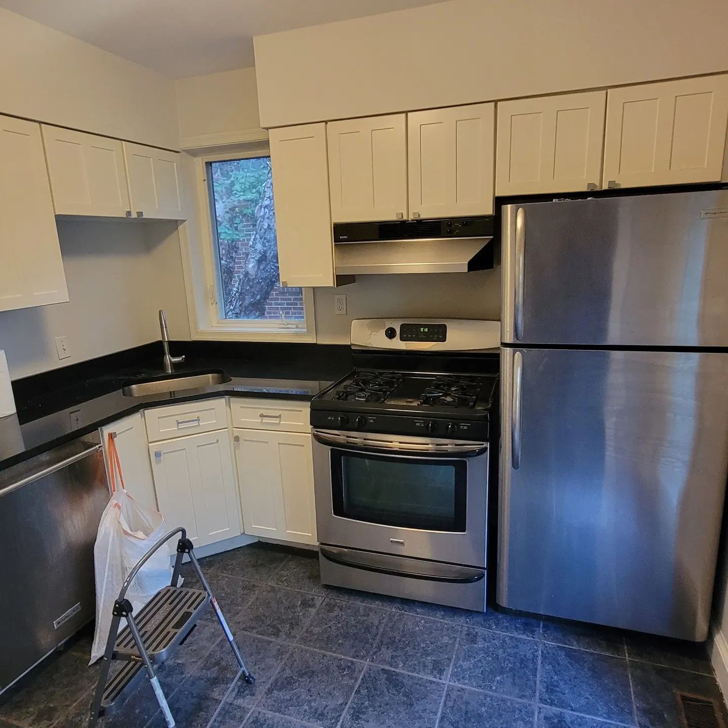 Small kitchen with white cabinets, black countertops, stainless steel appliances, and a step stool by the sink.