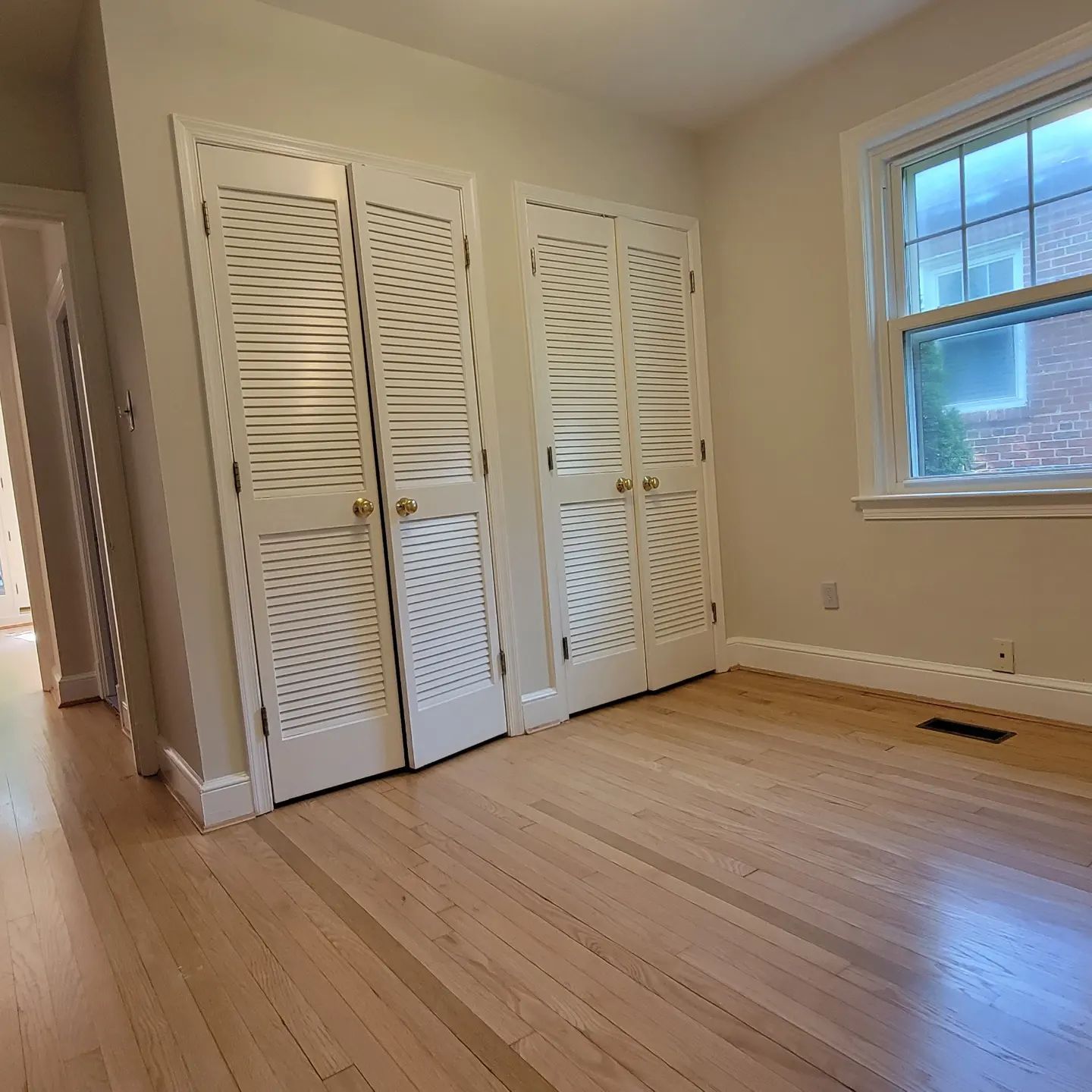 Empty room with hardwood floors, white louvered closet doors, and a window letting in daylight