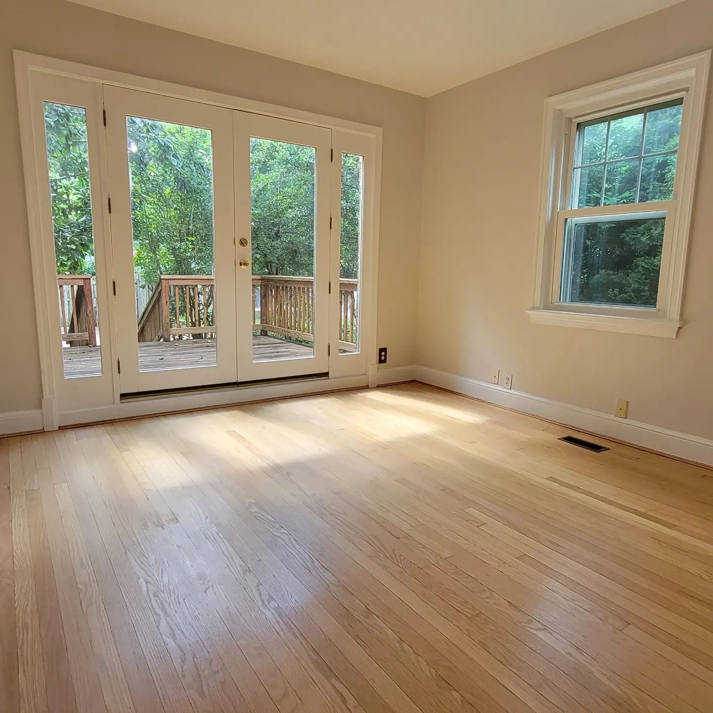 Empty sunlit room with hardwood floors, French doors to a deck, and a window overlooking trees.
