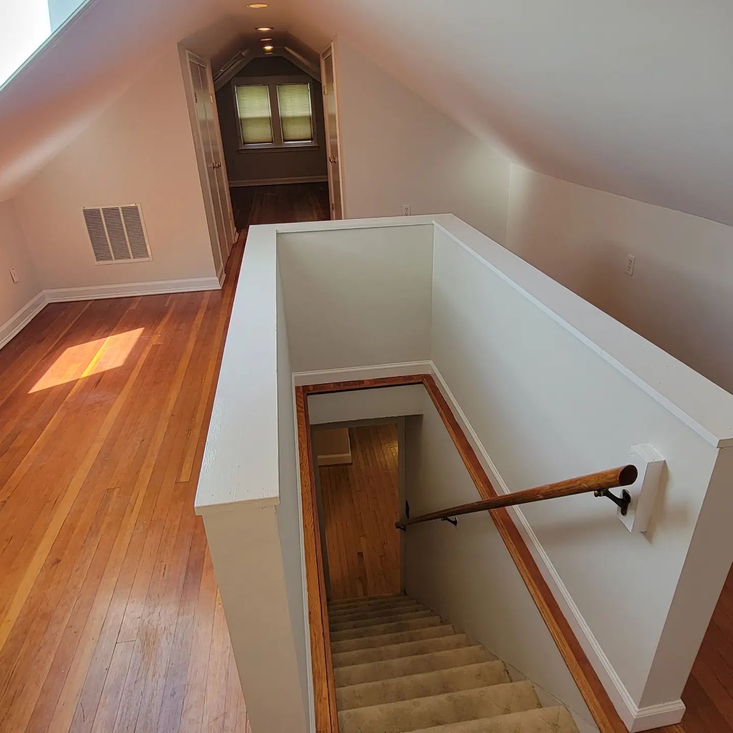 Attic hallway with wooden floor and open staircase railing leading down to a carpeted stairway.