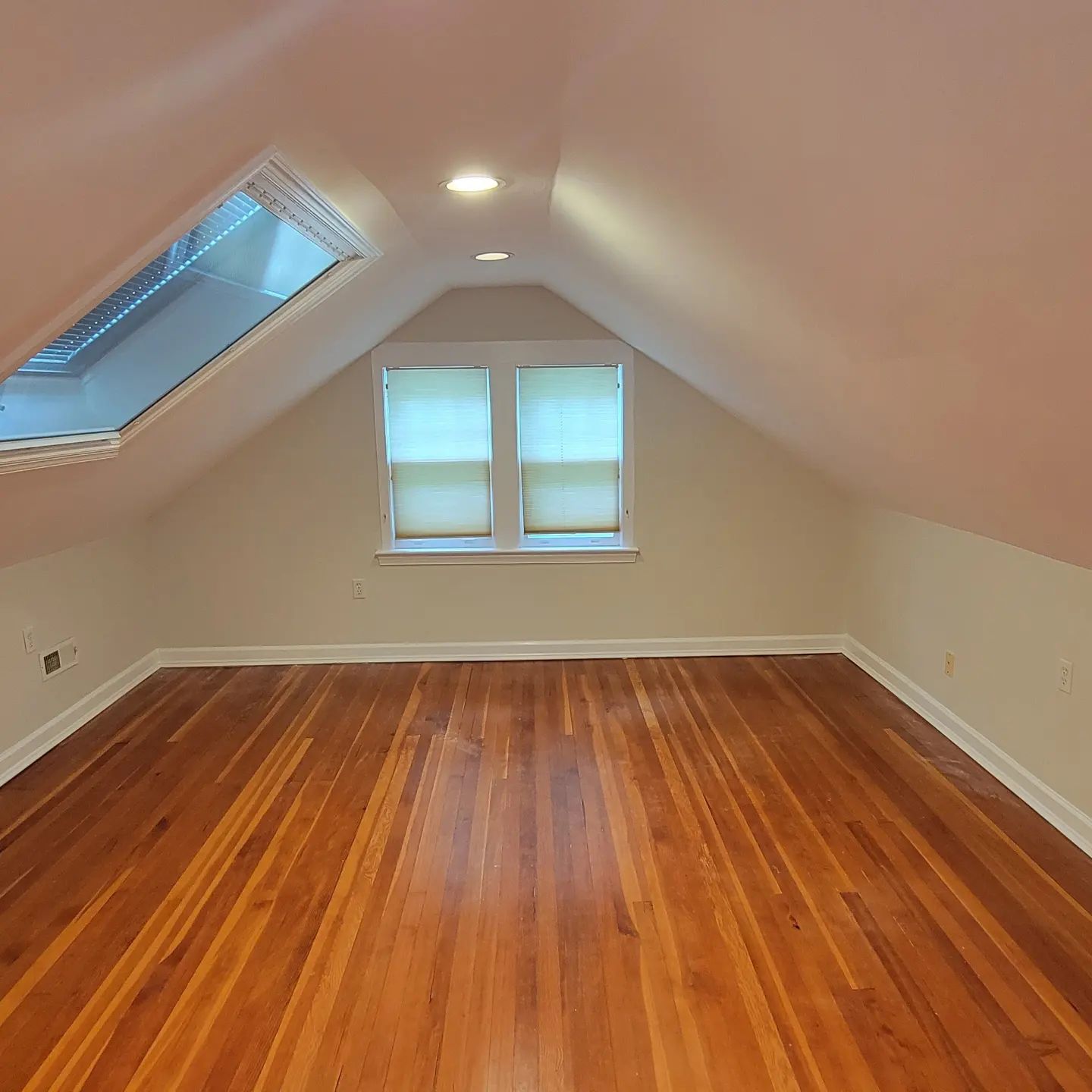 Empty attic room with hardwood floors, sloped white walls, skylight, and two small windows.