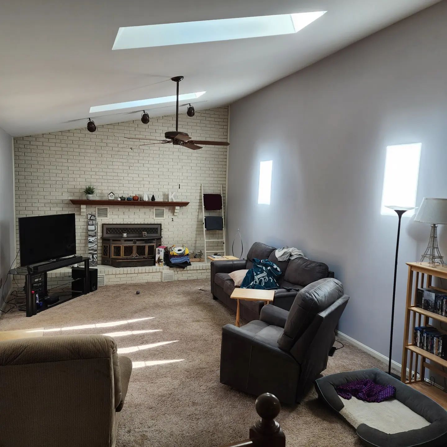 Living room with white brick fireplace, skylight, gray chairs, TV, and bright sunlight on carpet.
