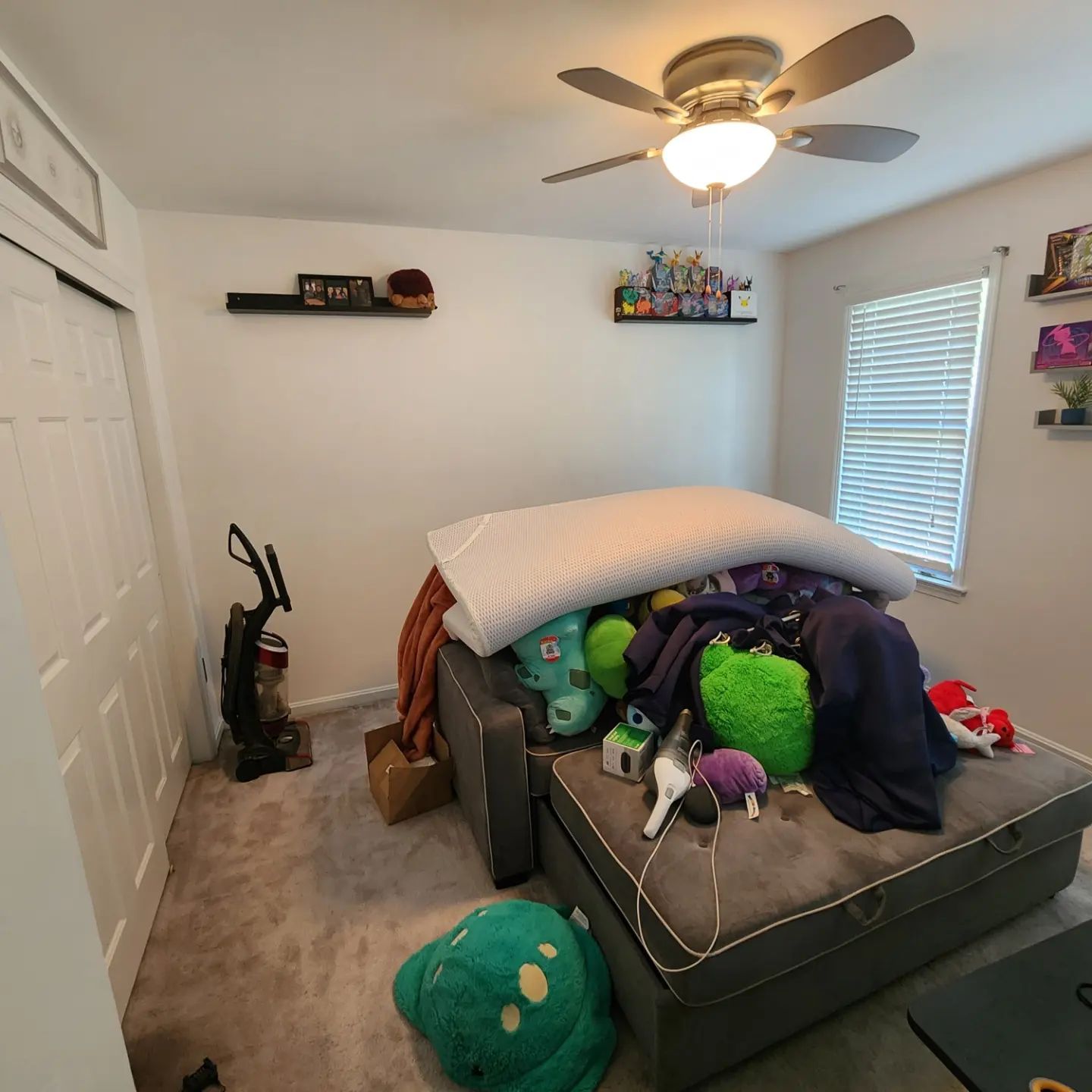 Small bedroom with a bed covered in pillows and stuffed animals, carpeted floor, and a ceiling fan.