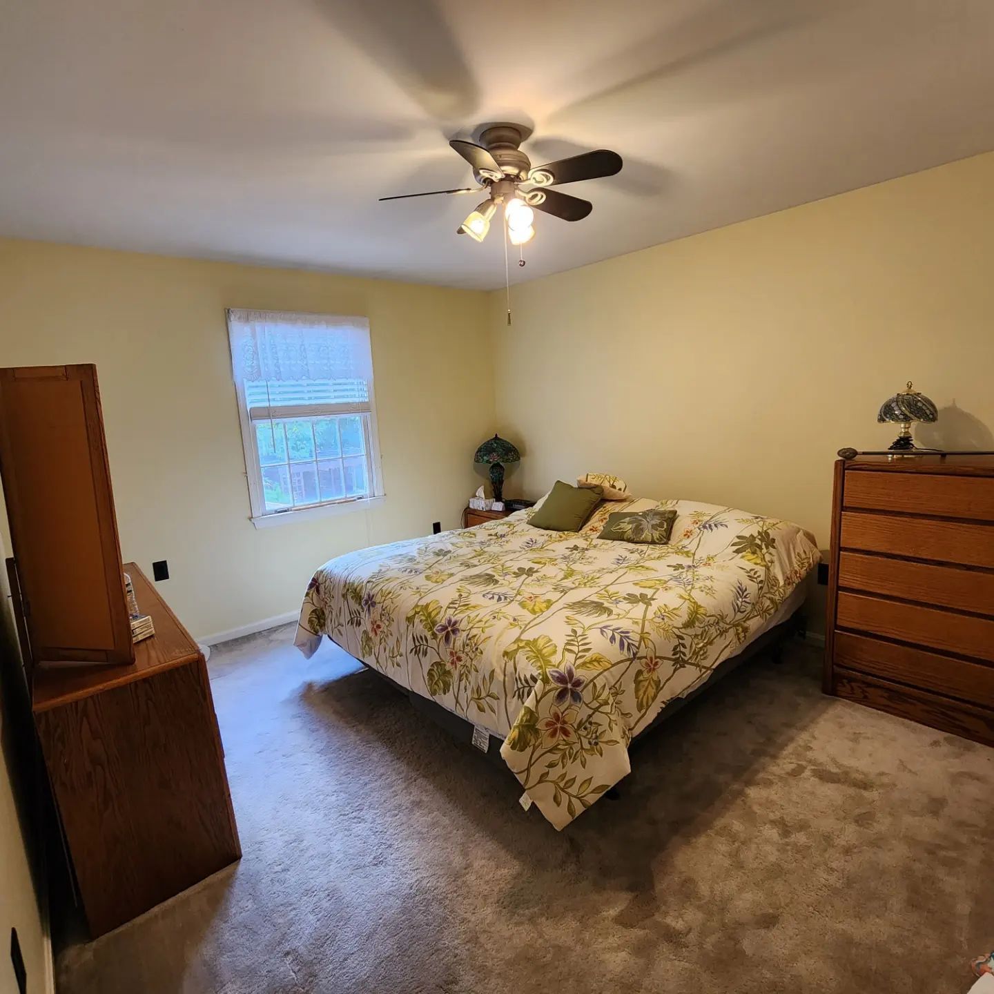 A tidy bedroom with a bed, dresser, window, ceiling fan, and beige carpet.