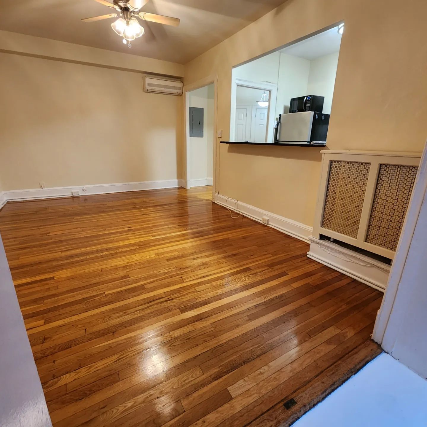 Empty living room with polished hardwood floors, beige walls, ceiling fan, and a pass-through kitchen opening.