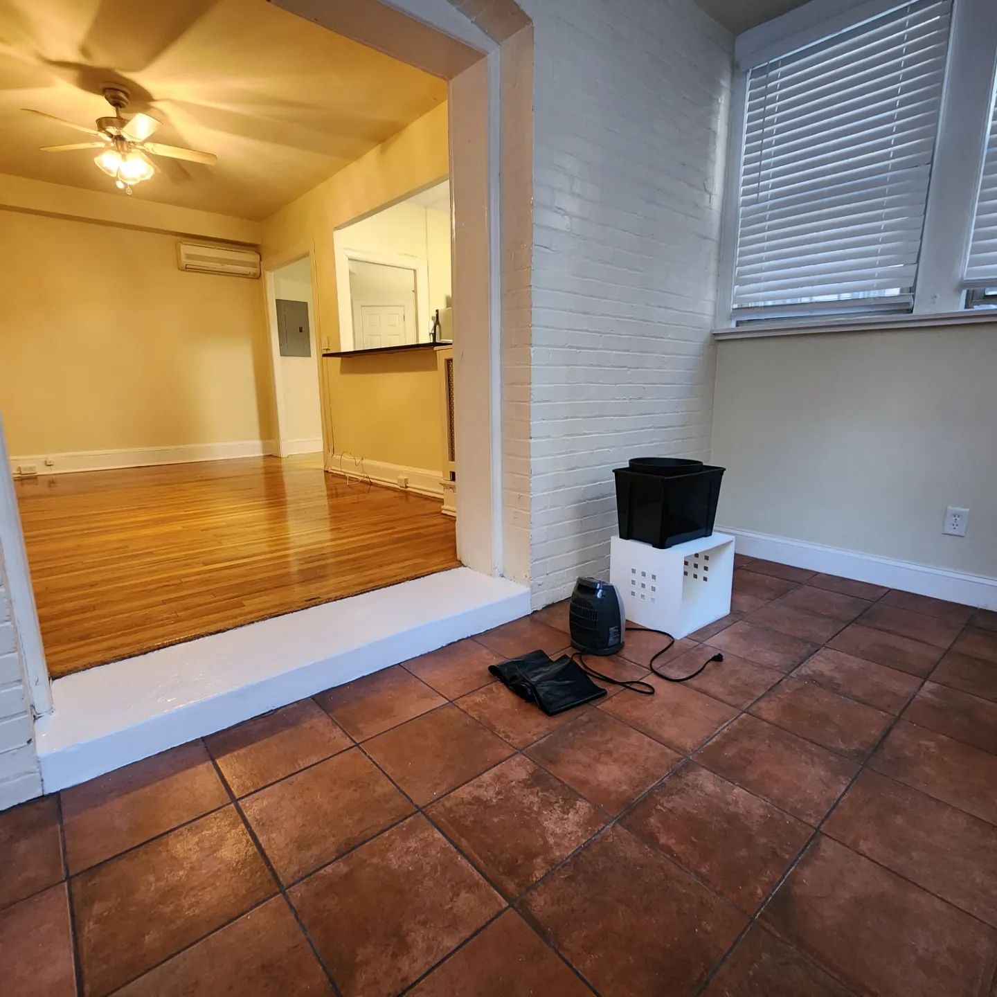 Empty tiled patio beside a living room with hardwood floors, window blinds, and a small pet crate and bowl.