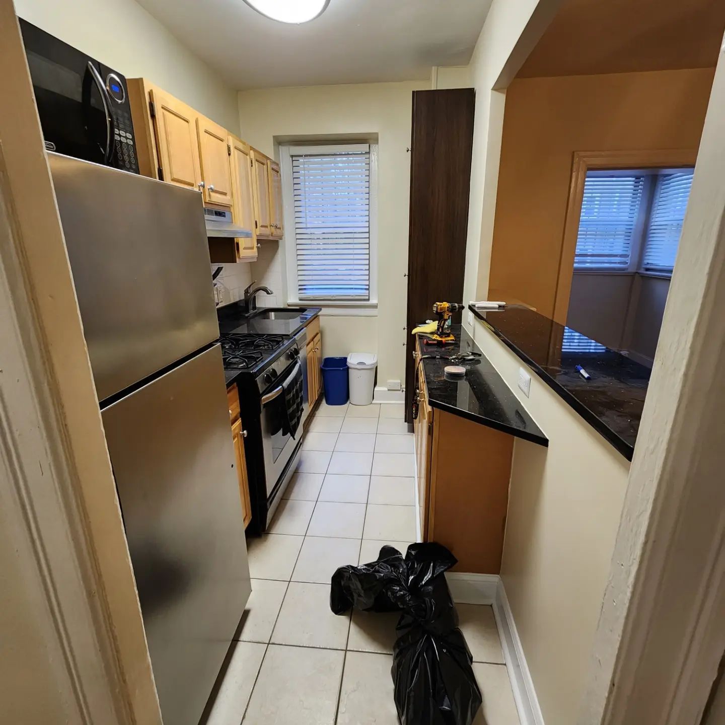 Narrow kitchen with stainless fridge, stove, wood cabinets, and black trash bags on the tiled floor.
