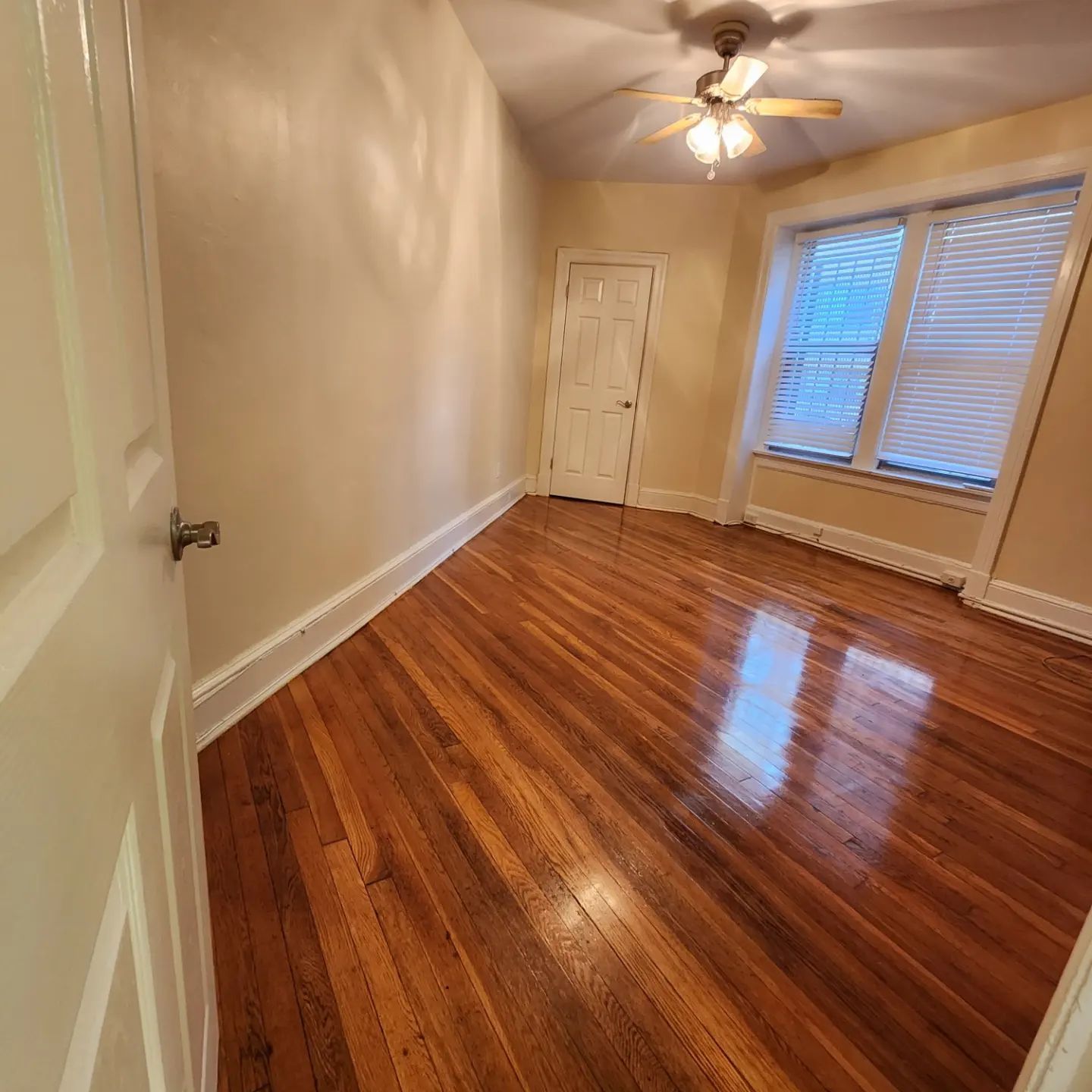 Empty room with polished hardwood floors, beige walls, ceiling fan, and windows with blinds.
