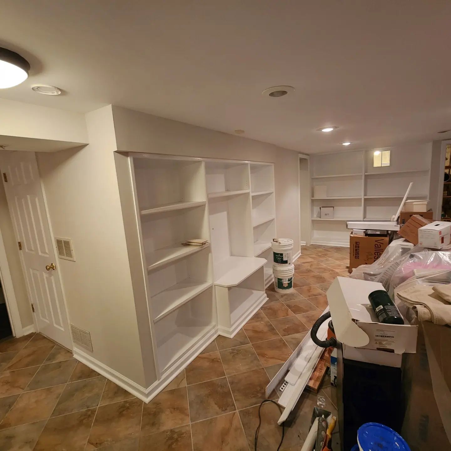 Unfinished basement with white built-in shelves, tiled floor, and renovation supplies scattered around