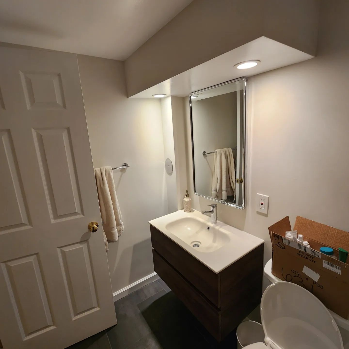 Small bathroom with sink, mirror, toilet, and open door; dark vanity and towel rack on left.