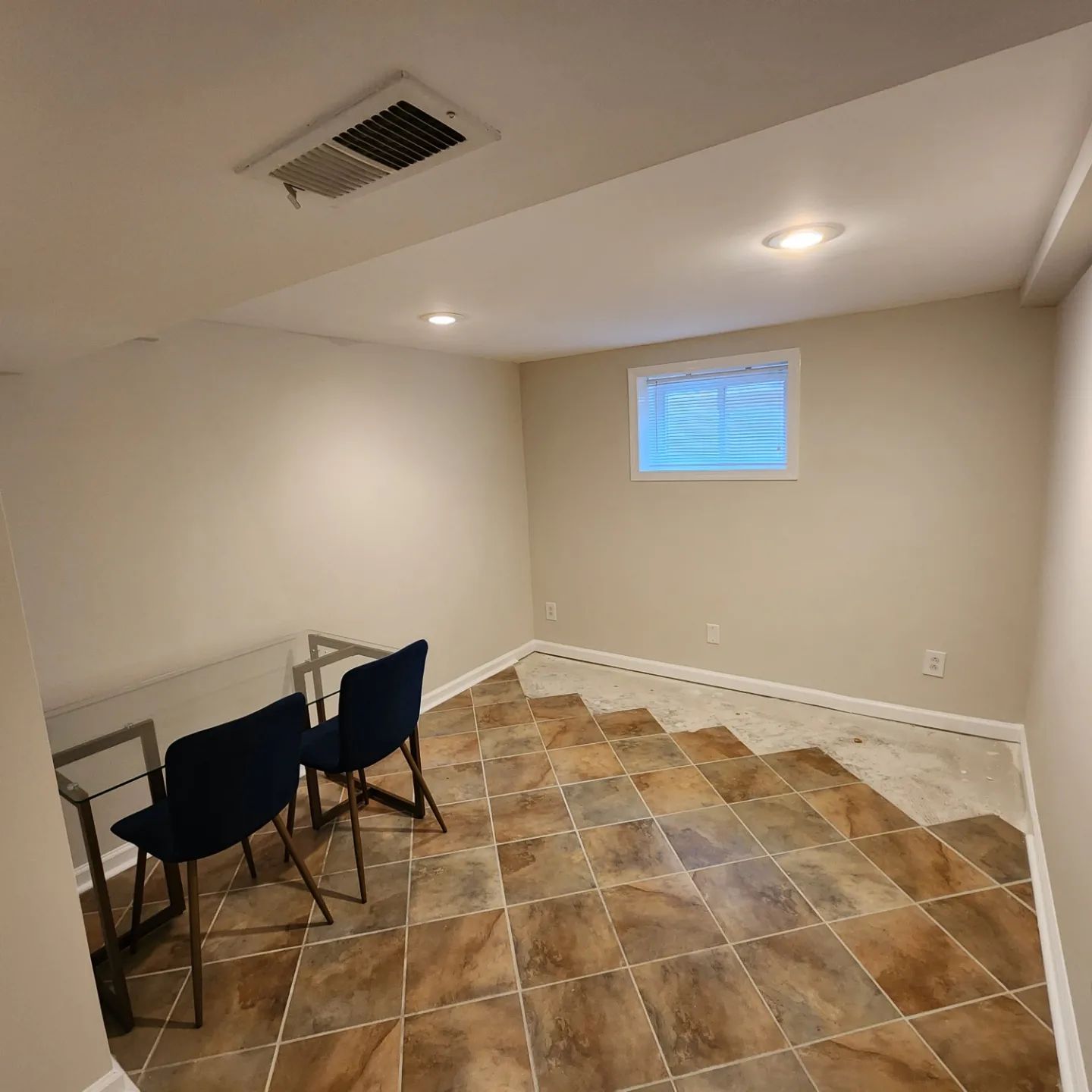 Empty basement room with tiled floor, white walls, small window, and two chairs by a table.
