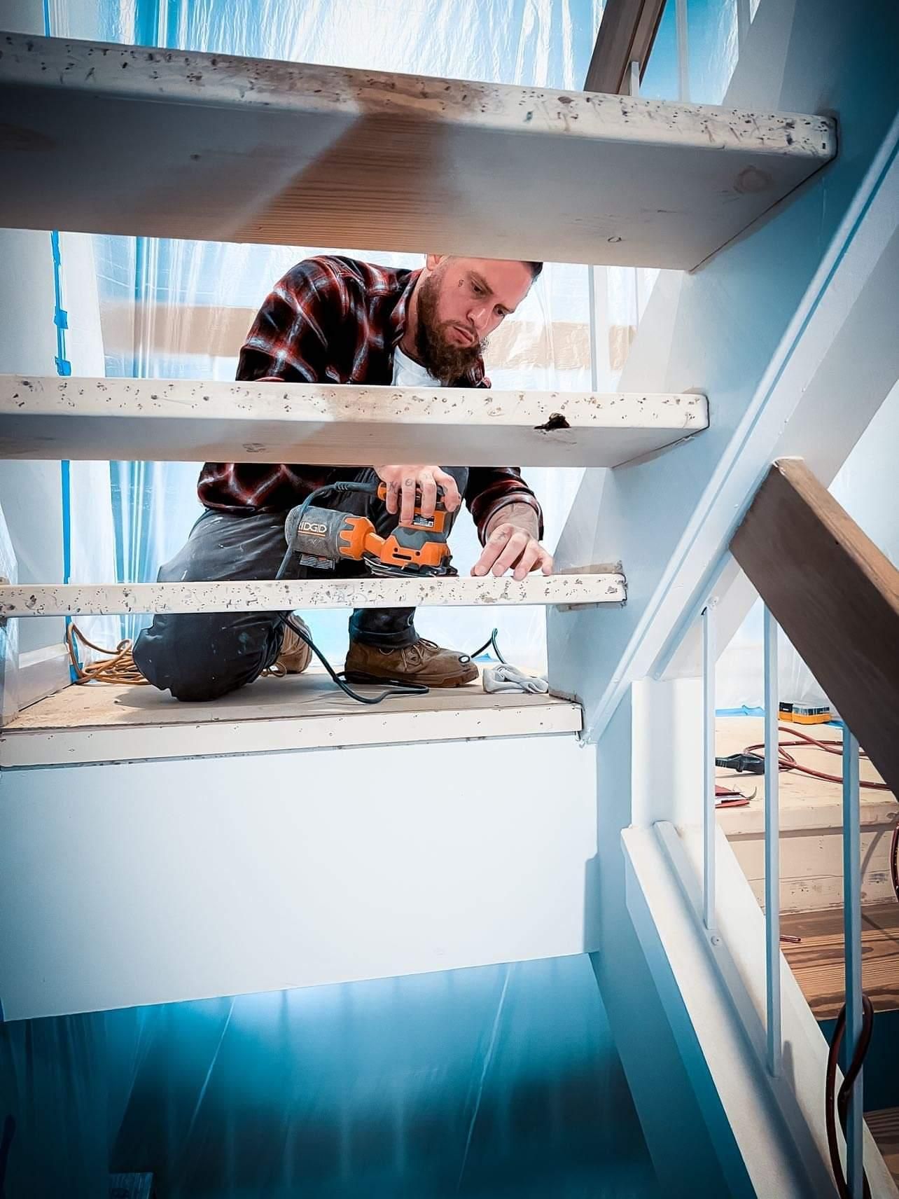 Worker installing wooden stairs in a bright interior, holding a drill beneath the steps