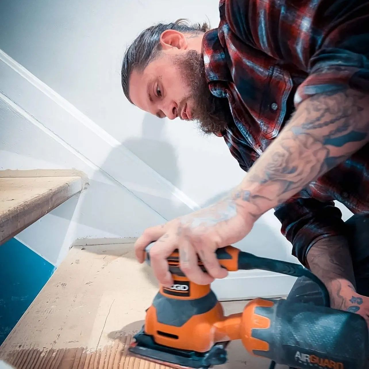 Man sanding a wooden surface with an orange power sander indoors, viewed from a low angle