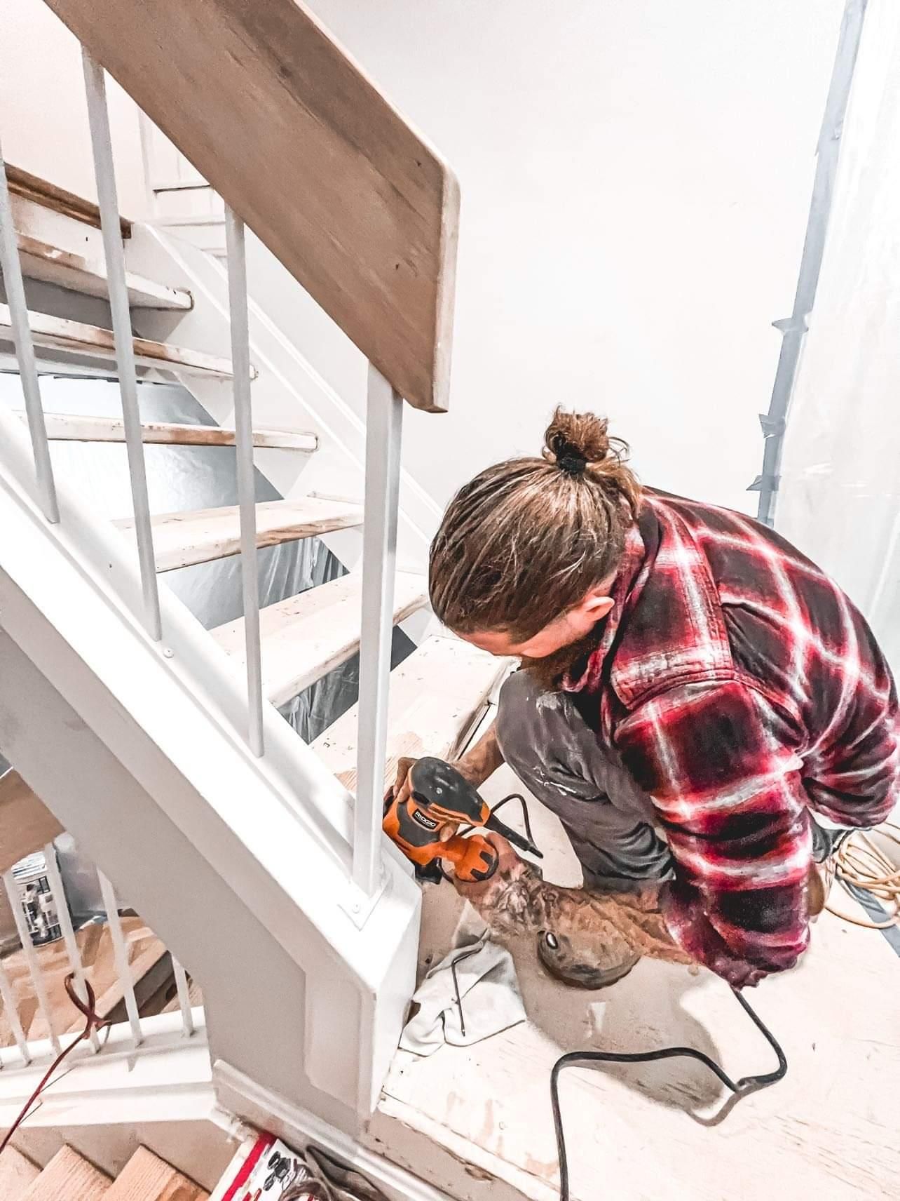 Person working with a power tool on a staircase landing, wearing a red plaid shirt.