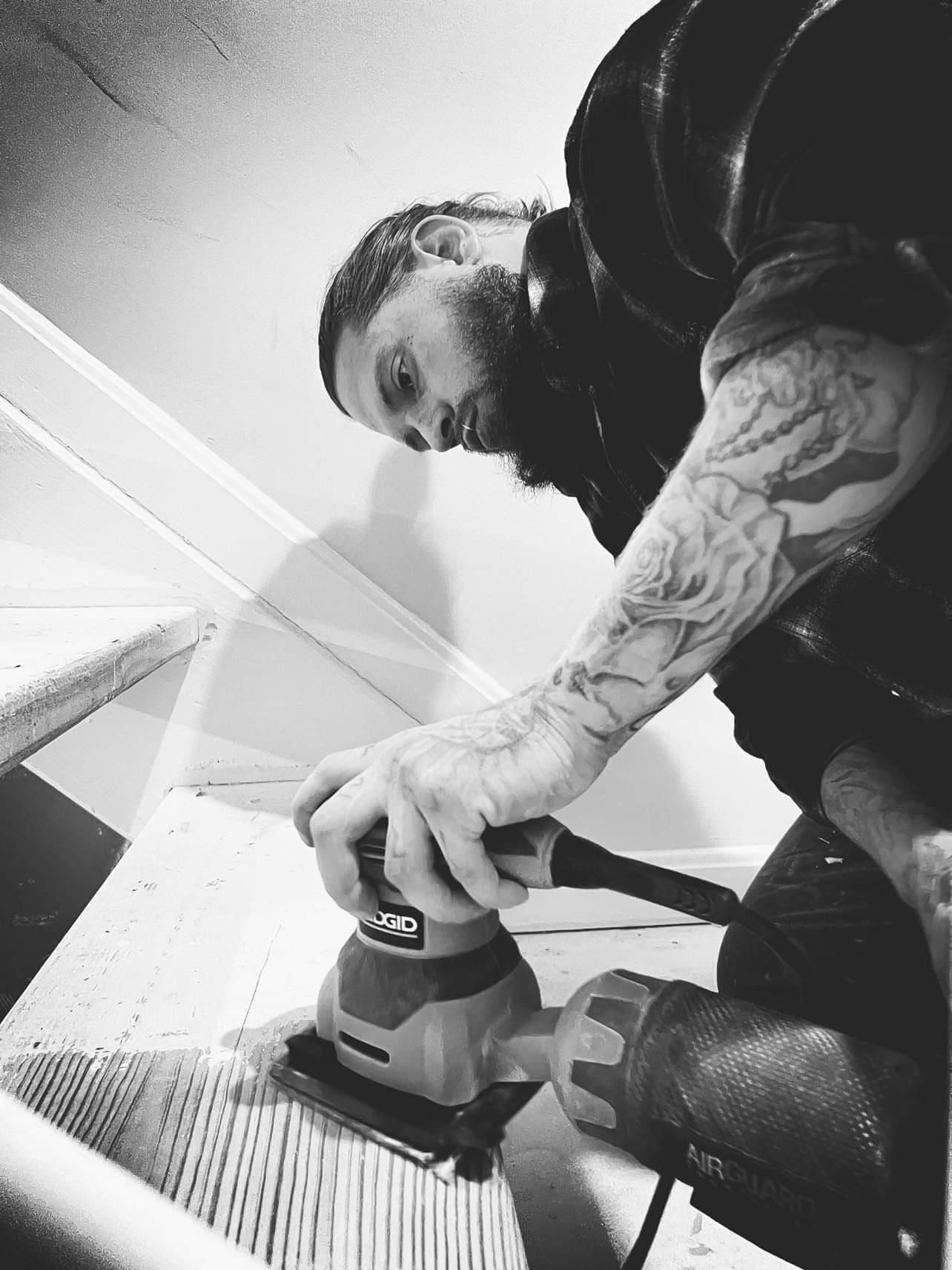 Person using a handheld sander on a wooden surface, viewed from below in black and white.
