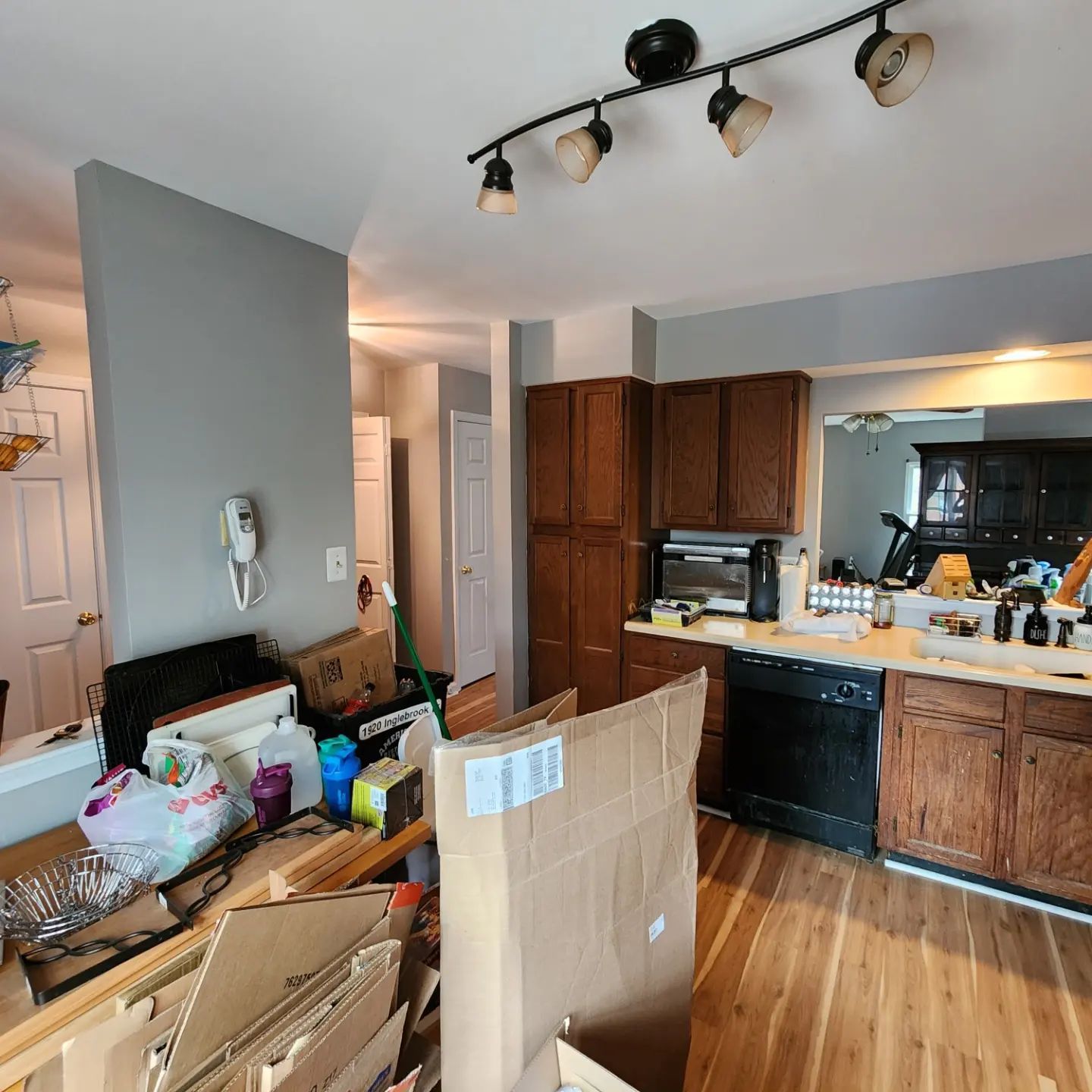 Messy kitchen with stacked cardboard boxes, wood cabinets, and black track lighting