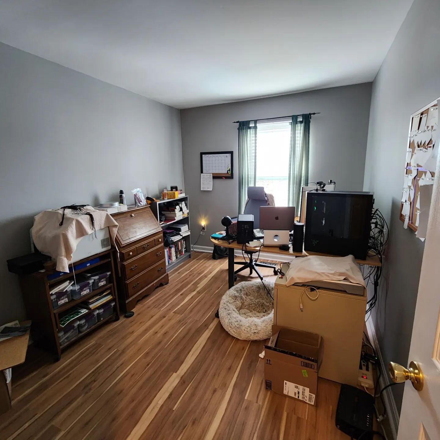 Cluttered bedroom with gray walls, wooden floor, dresser, TV, desk, and boxes near a window.