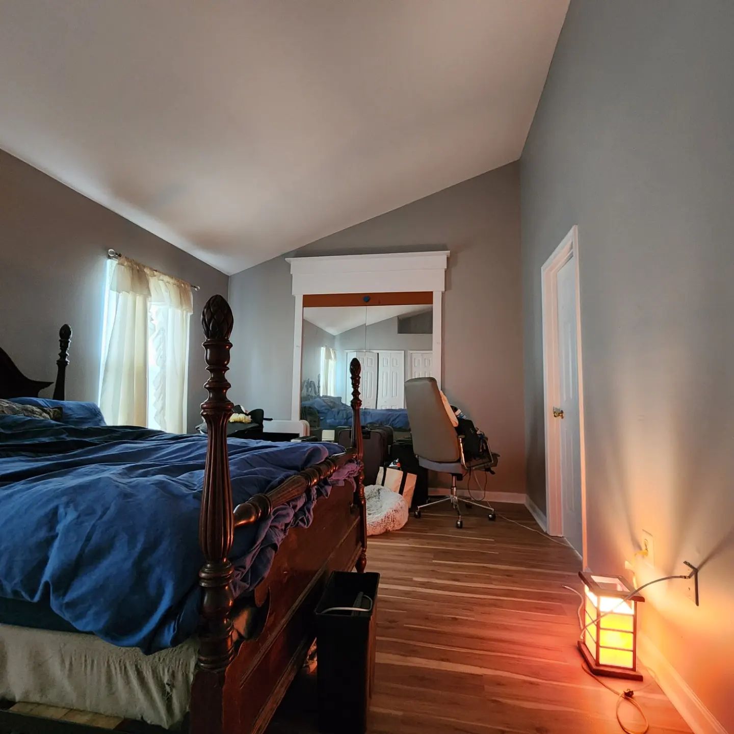 Bedroom with dark wood bed, desk by a window, warm lamp, and mirrored doorway reflecting the room.