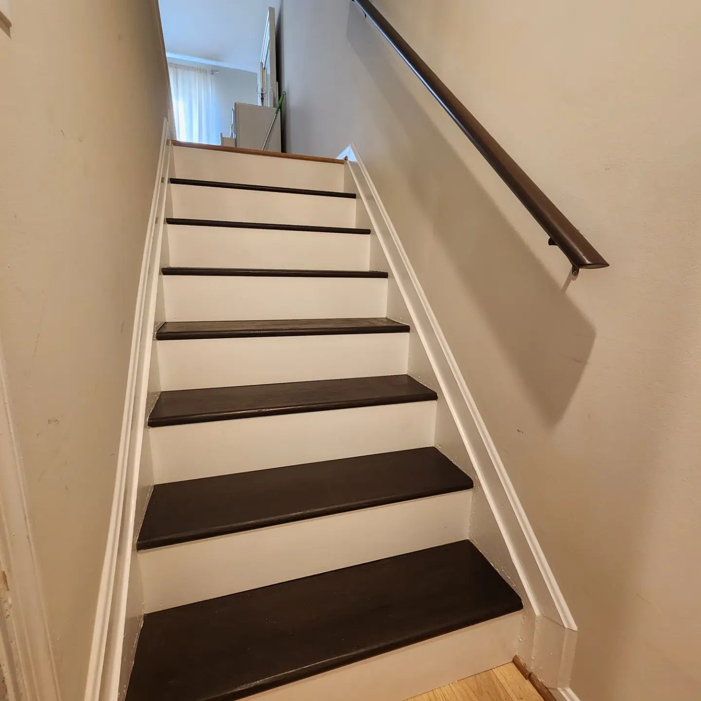 Narrow staircase with dark treads, beige walls, and a handrail leading to a bright landing