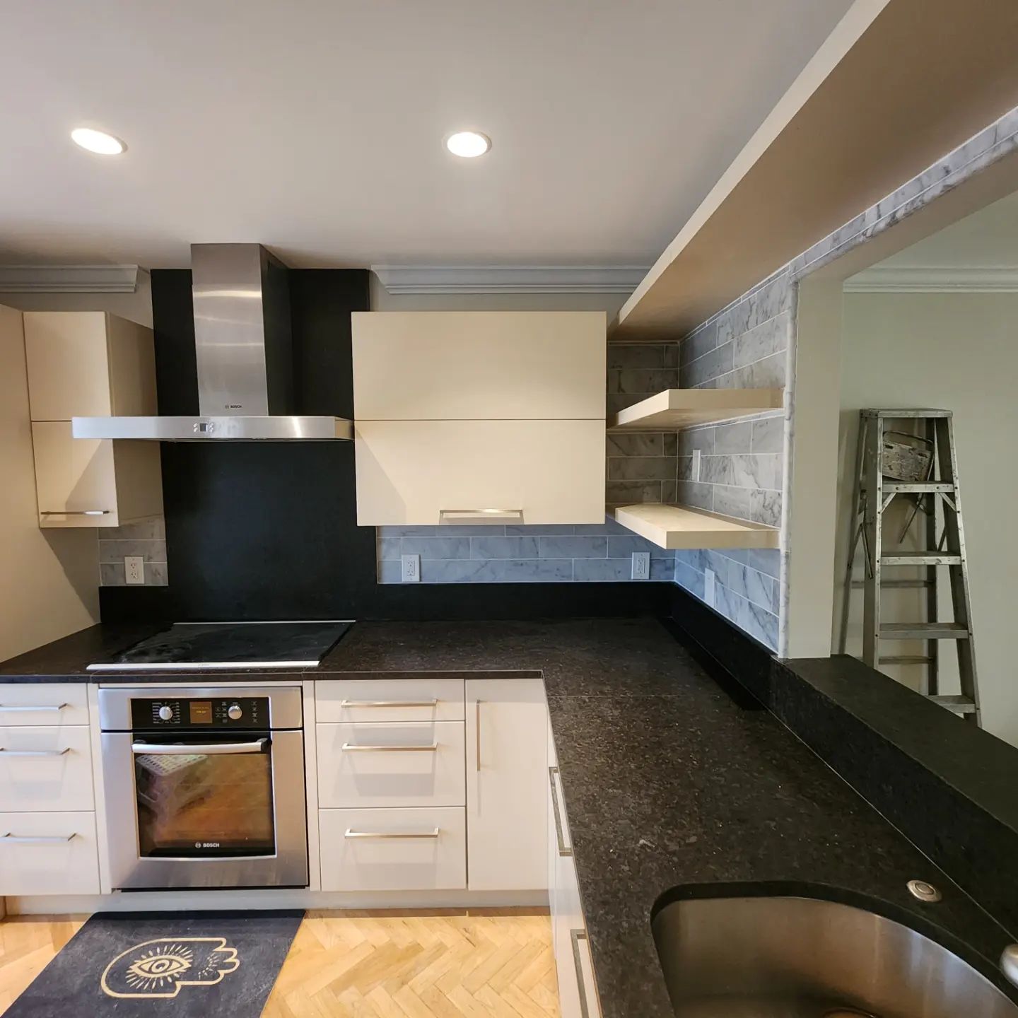 Modern kitchen with white cabinets, black countertops, stainless oven, and a sink by a reflective wall.