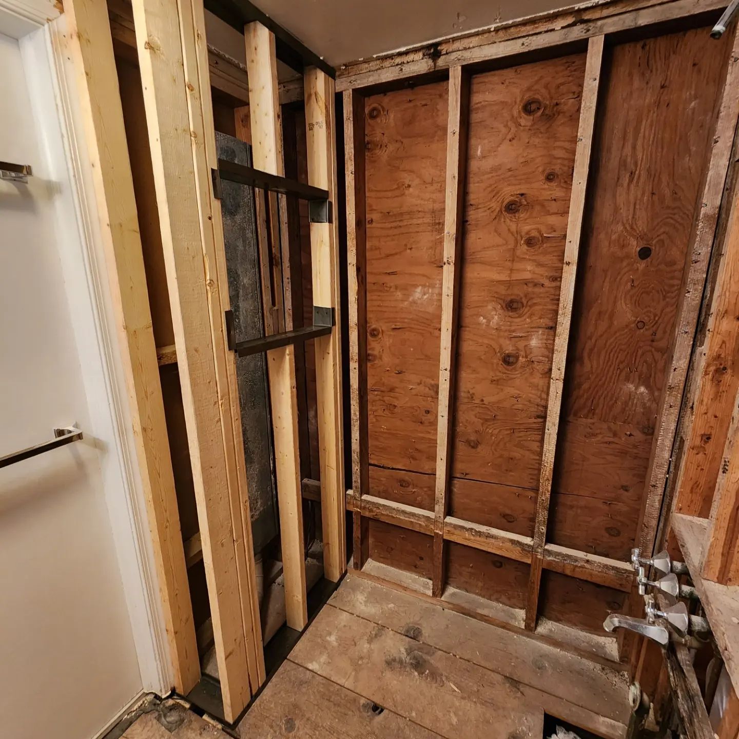 Exposed bathroom wall studs and plywood during renovation, with a doorway on the left.