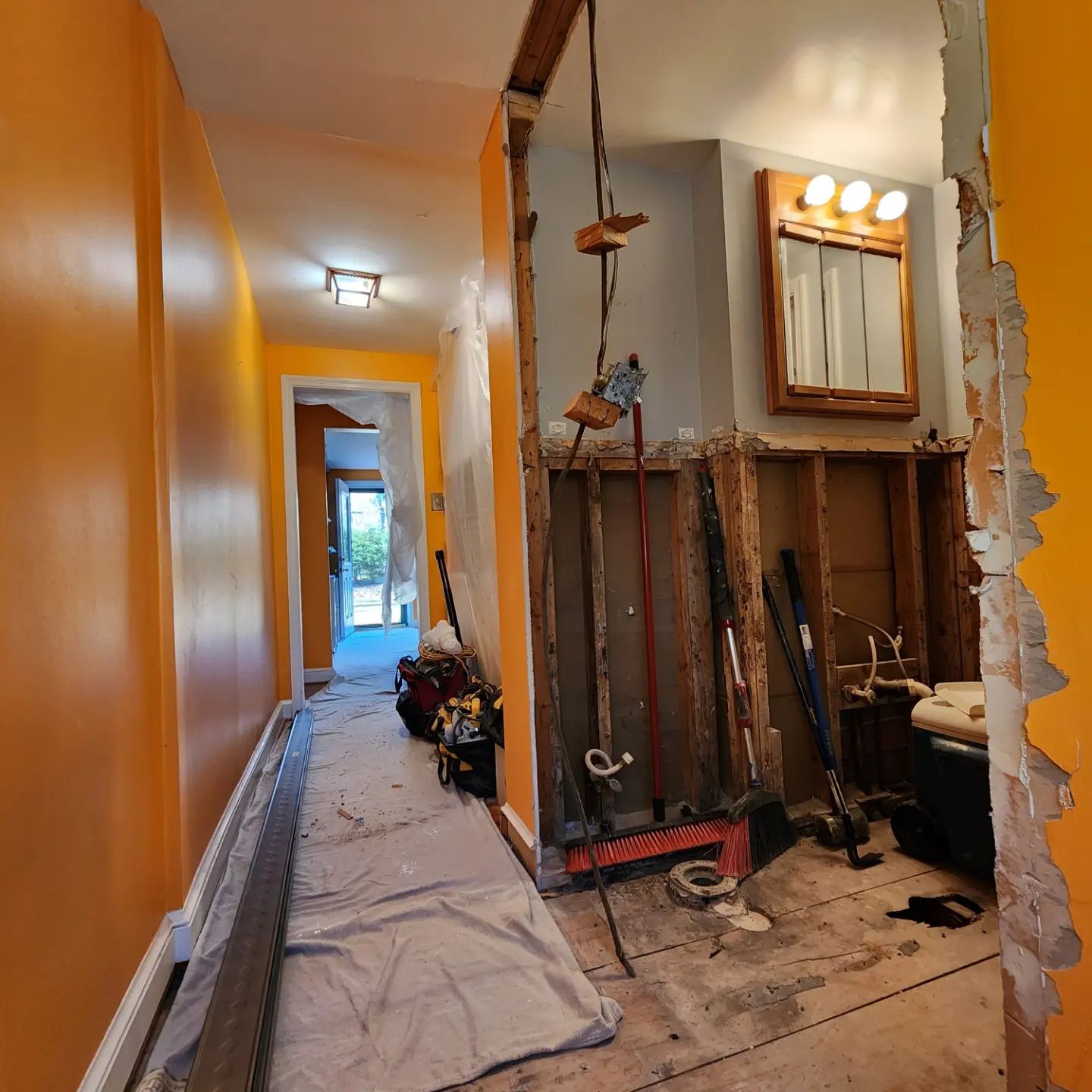 Narrow hallway under renovation with orange walls, exposed tools, and a lit bathroom mirror.