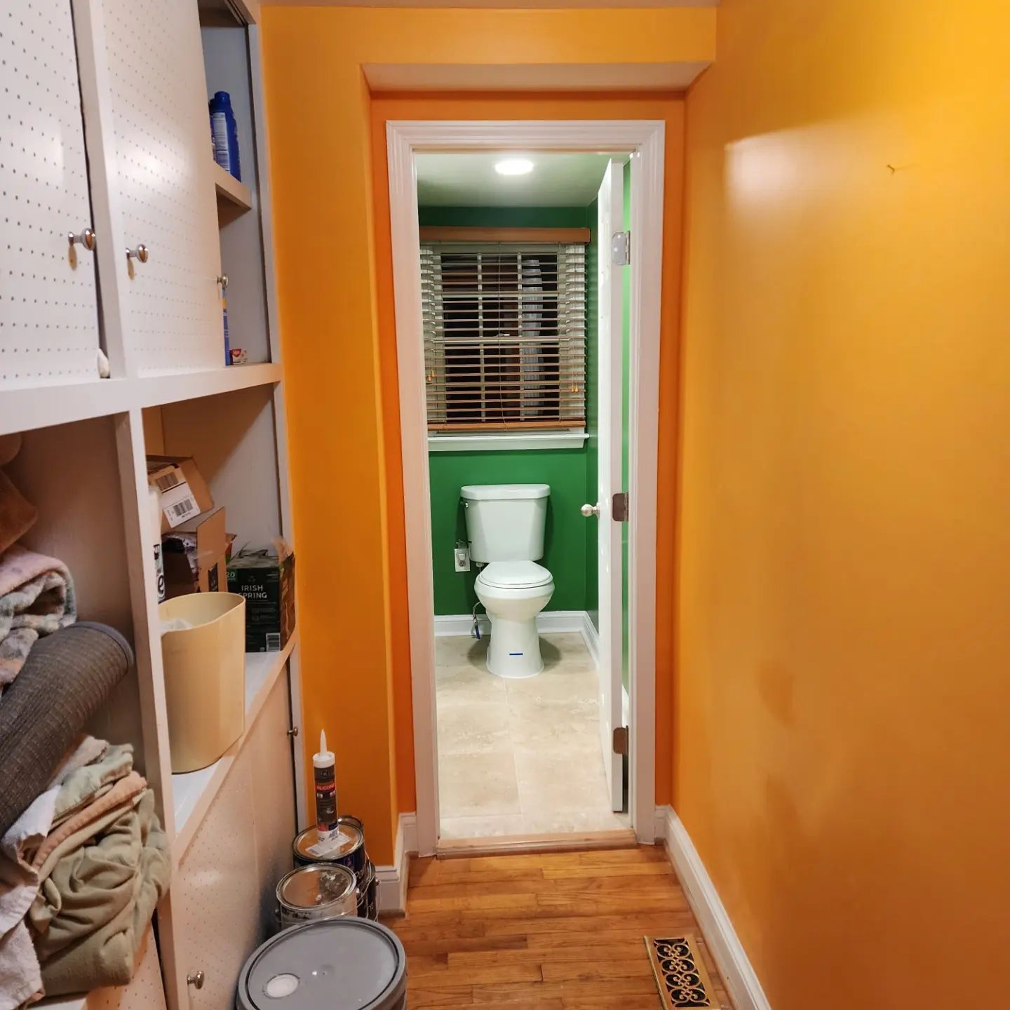 Narrow orange hallway leading to a green bathroom with a white toilet and window blinds