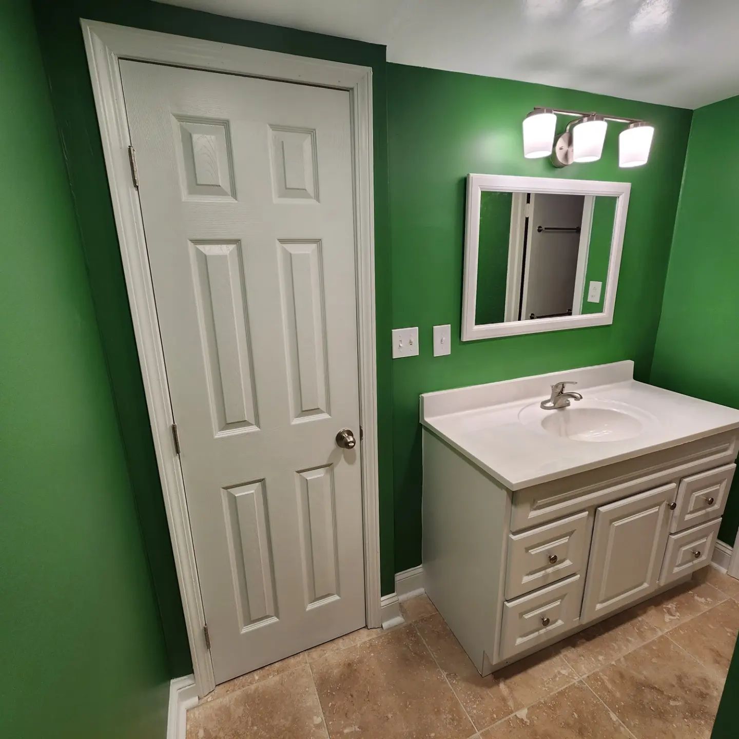 Small bathroom with green walls, white door, vanity sink, mirror, and light wood floor.