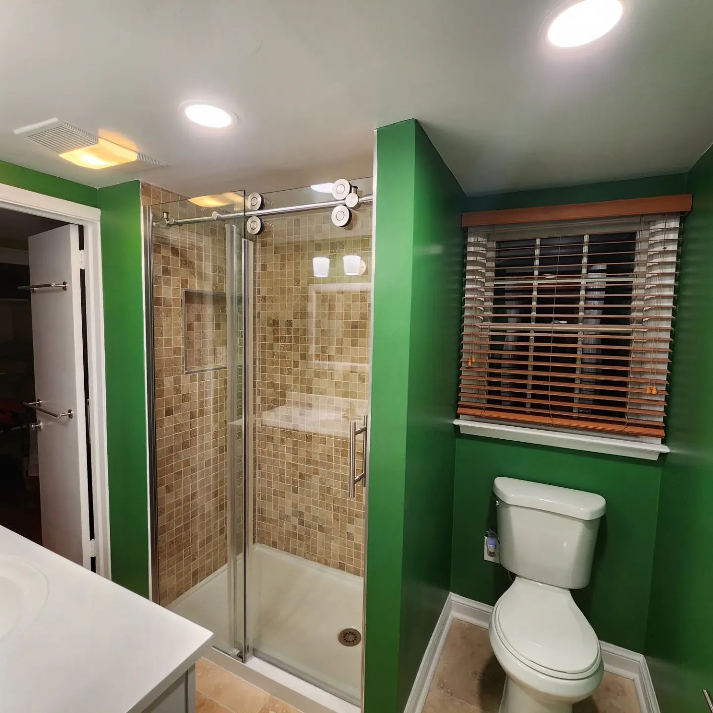 Small bathroom with green walls, shower stall, toilet, vanity, and window blinds.