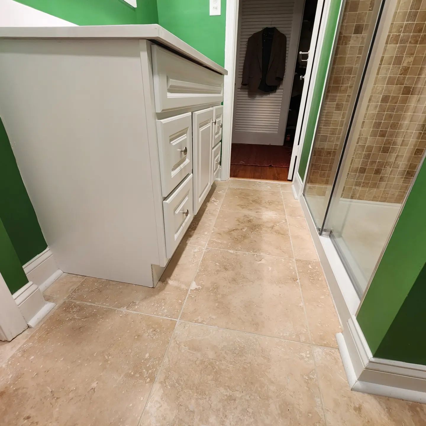 Narrow bathroom walkway with beige tile, green walls, white vanity on the left, and glass shower on the right