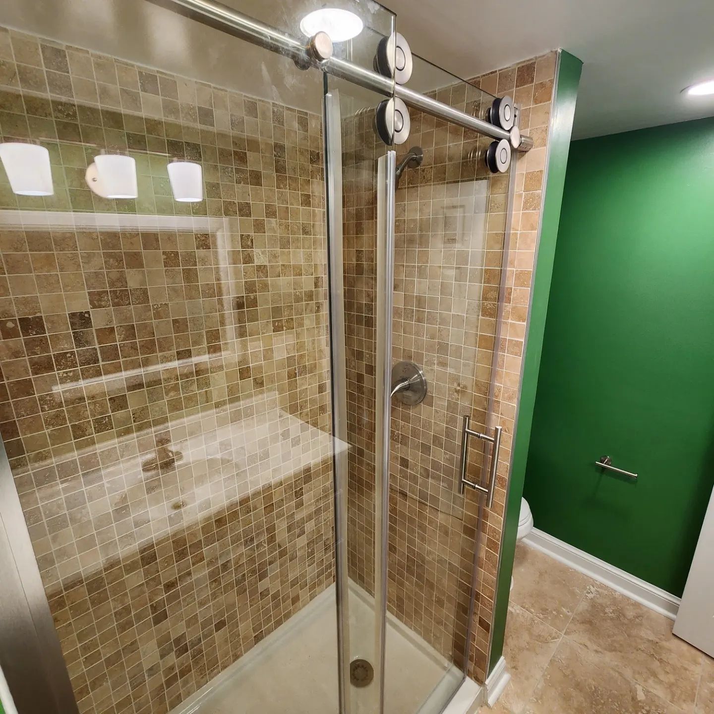 Glass shower enclosure in a beige-tiled bathroom with a green wall and wall-mounted shower bar.