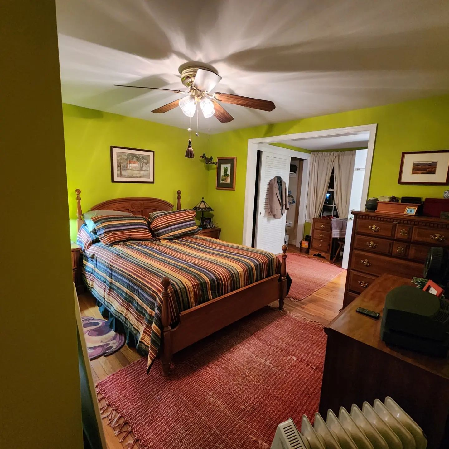 Bright bedroom with green walls, striped bedspread, ceiling fan, red rug, dresser, and open closet.