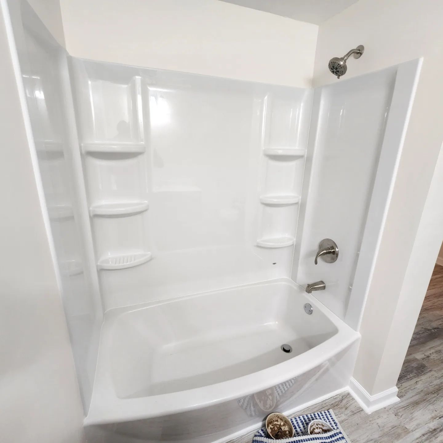 White bathtub and shower alcove with built-in corner shelves and chrome fixtures in a bathroom.