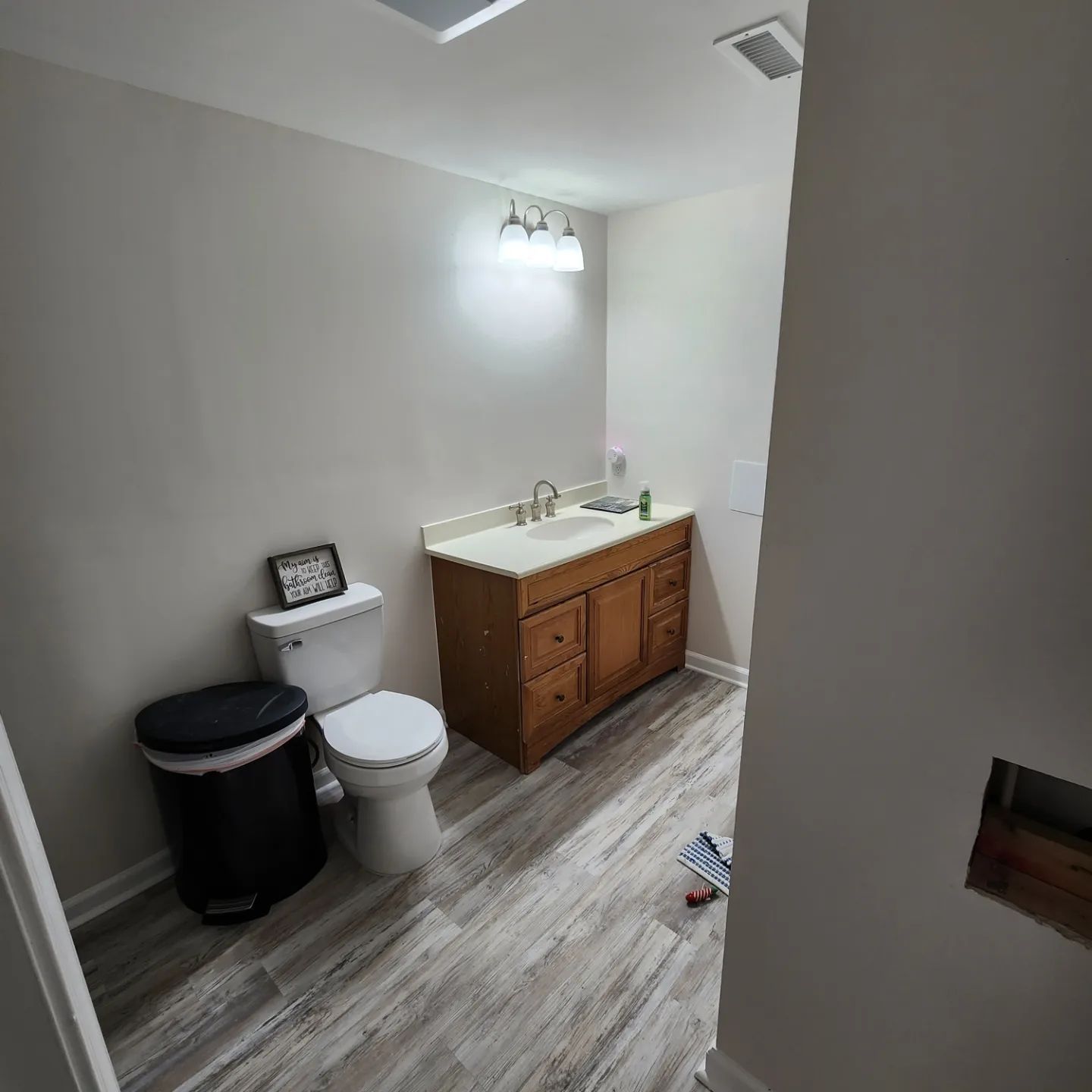 Small bathroom with toilet, dark trash can, wood vanity, sink, and wall light on gray walls.