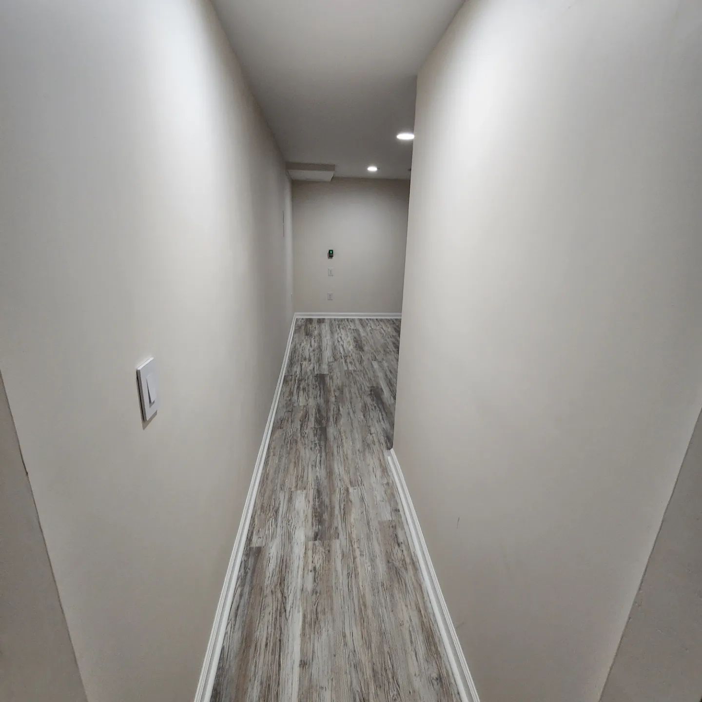 Narrow hallway with white walls, gray wood floor, recessed ceiling lights, and an open doorway at the end