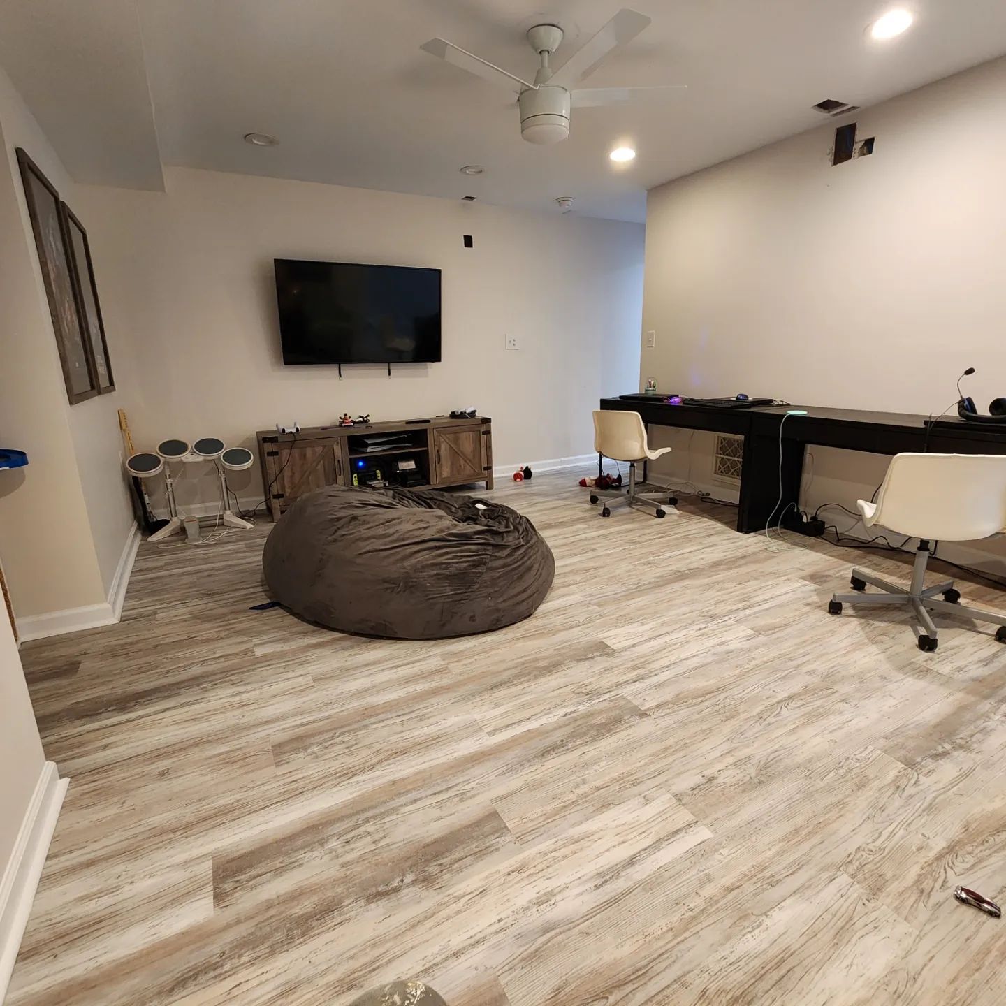 Spacious basement media room with TV, bean bag chair, desks, and white chairs on wood-look flooring