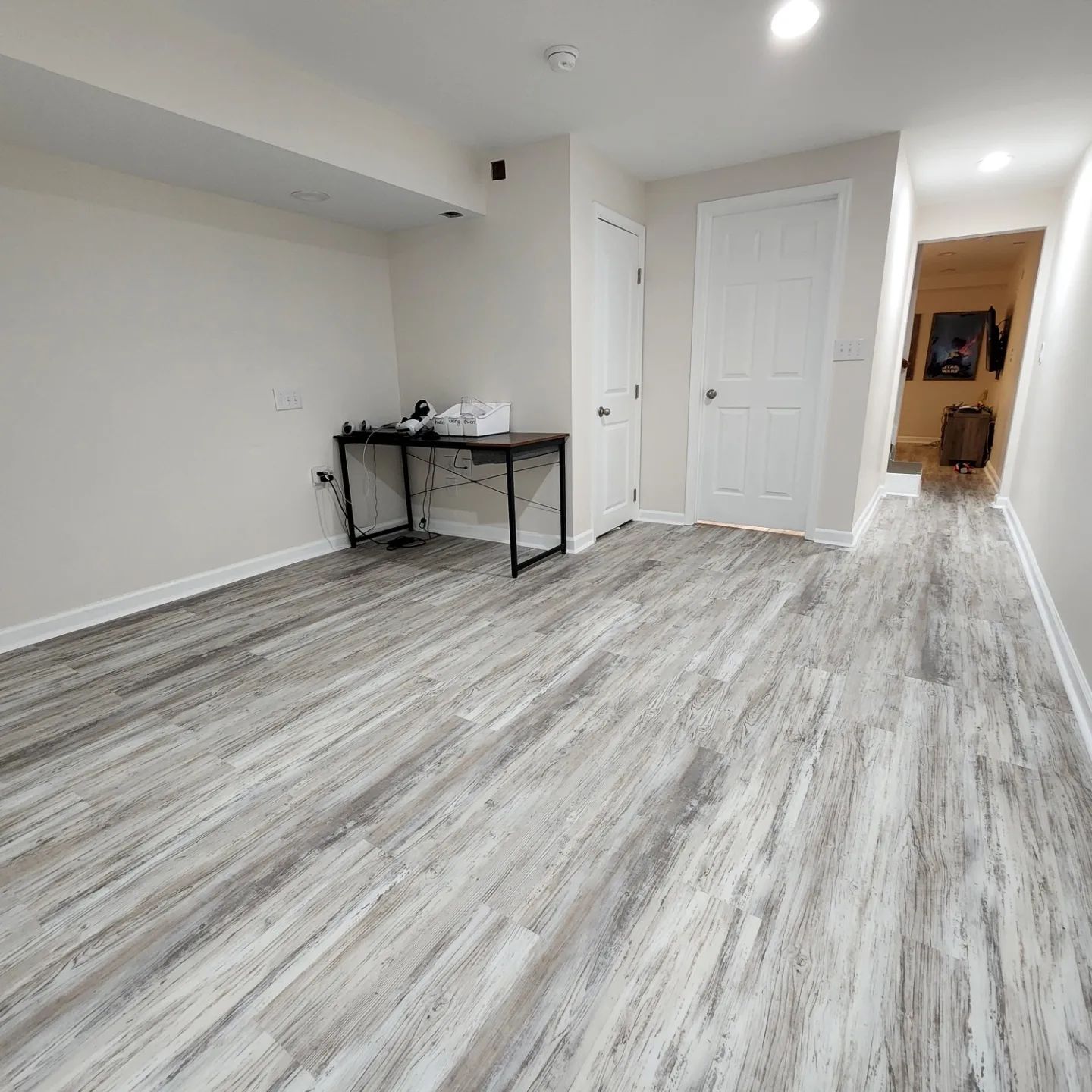 Empty white room with gray wood-look flooring, a black desk, and closed doors leading to a hallway.