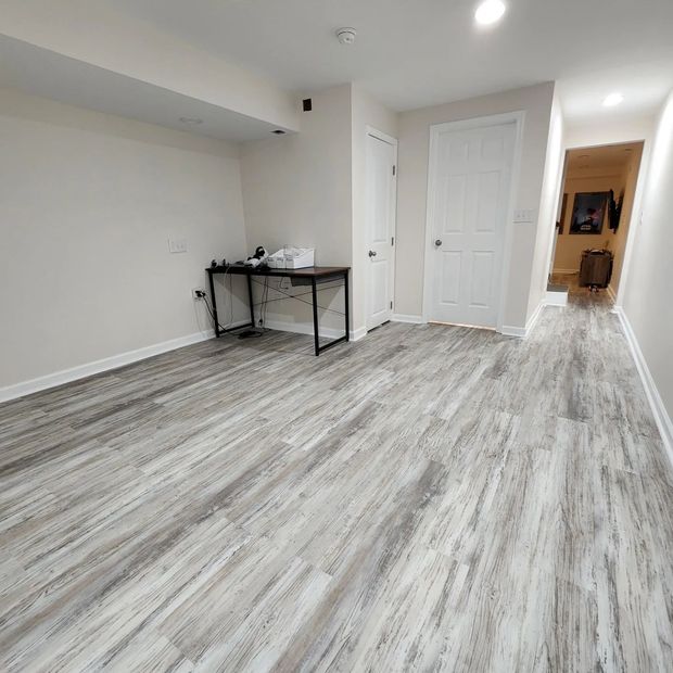 Empty room with gray wood flooring, white walls, a small desk, and a hallway leading to another room.